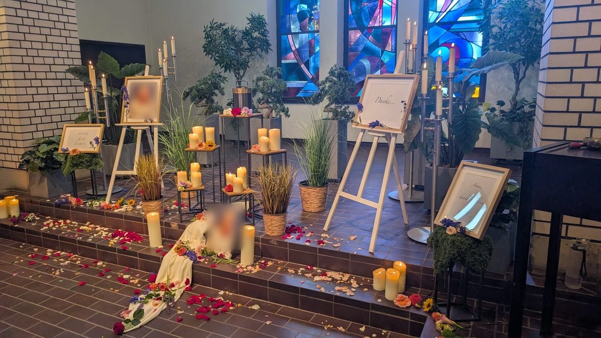 Gesamtansicht einer Trauerfeier in einer Kirche: Kerzen, Pflanzen und Blütenblätter auf den Stufen, Staffeleien mit Dankeskarten und dem unkenntlich gemachten Bild der Verstorbenen
